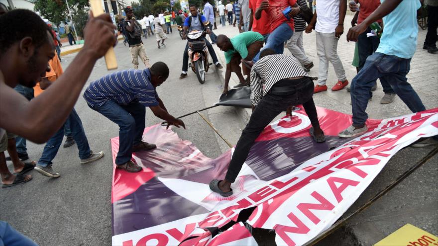 Manifestantes rompen un cartel electoral del candidato presidencial Jovenel Moise durante una marcha