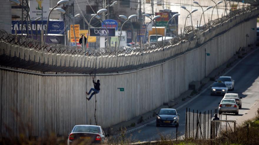 Un palestino intenta cruzar por encima de una sección del controvertido muro de separación israelí que separa la ciudad cisjordana de Al-Ram del este de Al-Quds (Jerusalén), 24 de febrero de 2016.