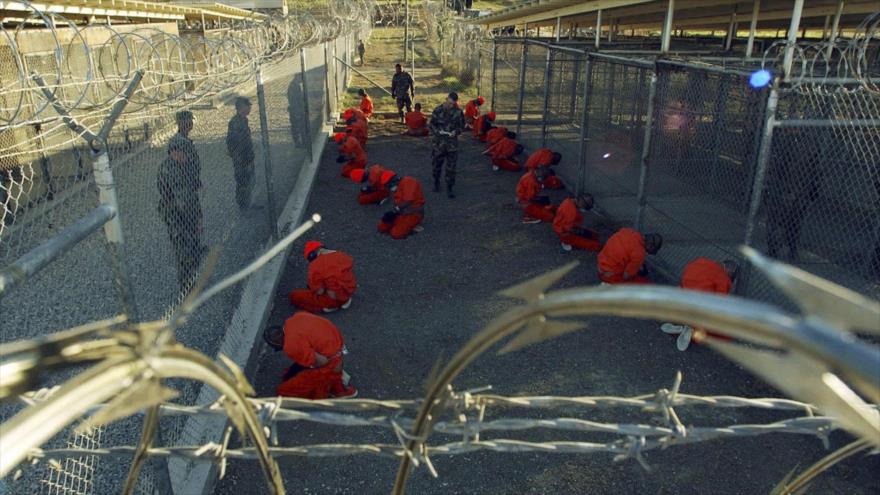Los detenidos en trajes de color naranja se sientan en una zona de espera bajo la mirada vigilante de la policía militar durante el procesamiento hasta el centro de detención temporal en el Campamento Rayos X de la base naval de Guantánamo, en Cuba, 11 de enero de 2002.
