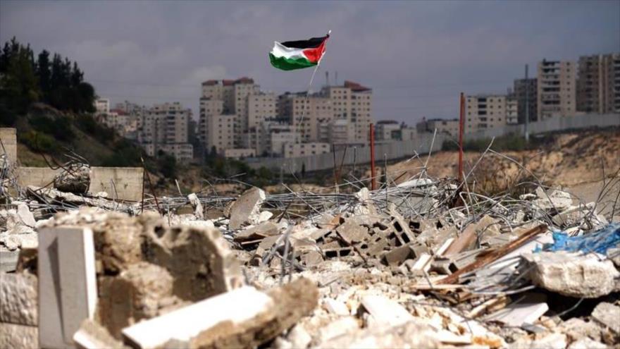 La bandera nacional de Palestina ondea sobre los escombros de casas palestinas destruidas por el régimen de Israel en Al-Quds (Jerusalén).