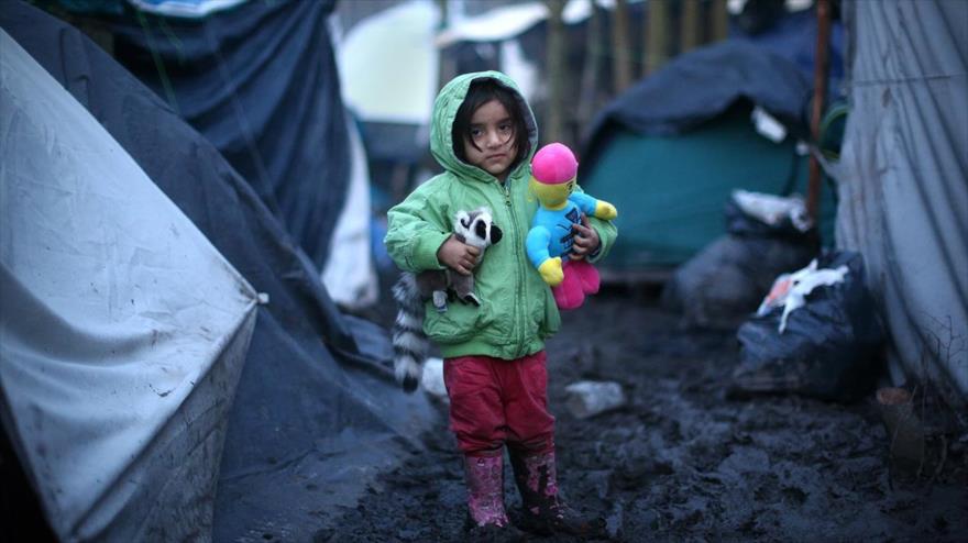 Una niña kurda sostiene sus juguetes mientras está en el barro en un campamento de refugiados en la costa norte de Francia.