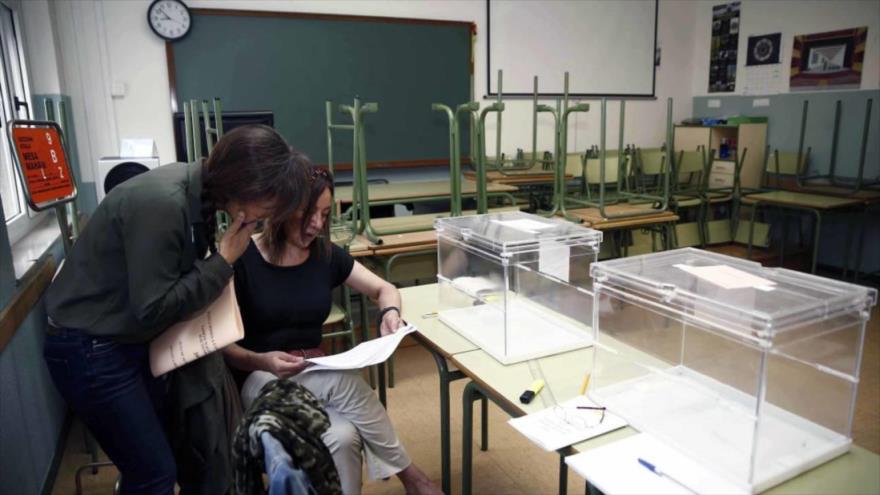 Dos mujeres revisan un listado electoral ante dos urnas vacías en un colegio electoral en España.