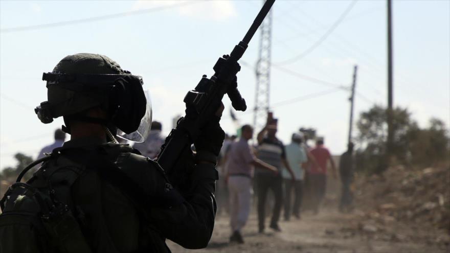 Un soldado israelí mantiene arriba su arma, dispuesto a disparar, mientras un grupo de palestinos se aleja del lugar.