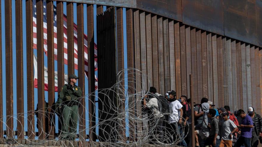 Migrantes centroamericanos miran a través de la valla mientras un agente vigila cerca del cruce fronterizo de El Chaparral en Tijuana, México.