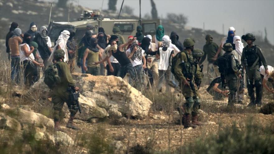 Soldados israelíes junto a colonos enmascarados que arrojan piedras a los manifestantes palestinos en Cisjordania, 17 de octubre de 2019. (Foto: AFP)