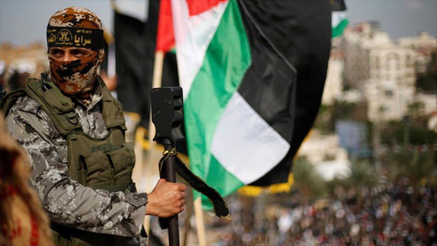 Un combatiente de Saraya Al-Quds, rama militar de la Yihad Islámica, sostiene su armamento, mientras la bandera palestina ondea en el fondo.
