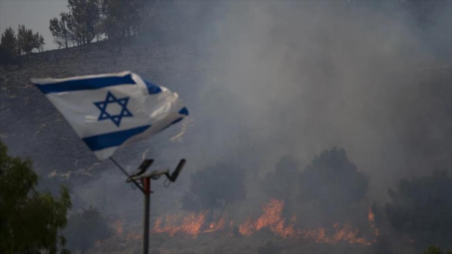 Una bandera israelí ondea en una zona que es arrasada por un incendio en Safed, el norte de Palestina ocupada, 12 de junio de 2024.