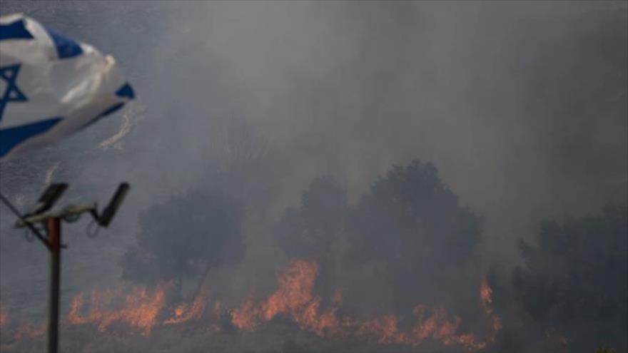 Una bandera israelí ondea en una zona que es arrasada por un incendio en Safed, el norte de Palestina ocupada, 12 de junio de 2024.