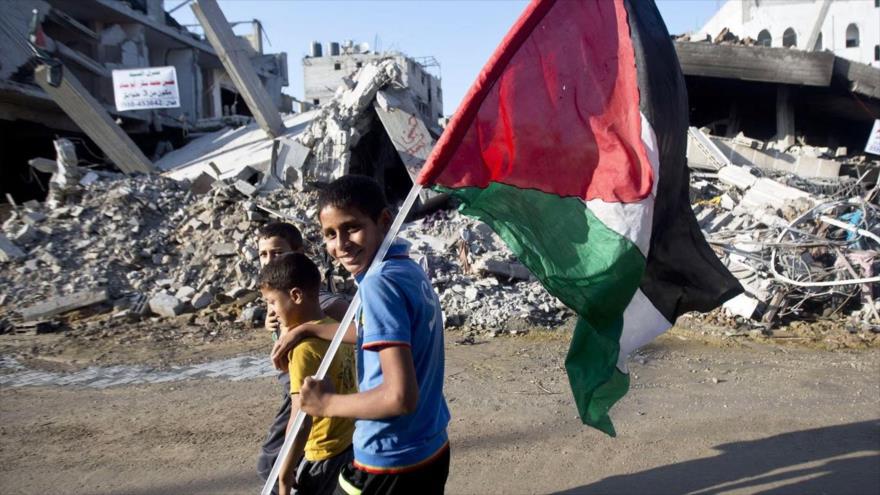 Niños palestinos sonríen y portan la bandera nacional mientras caminan por una escena de destrucción en la Franja de Gaza. (Foto: Getty Images)