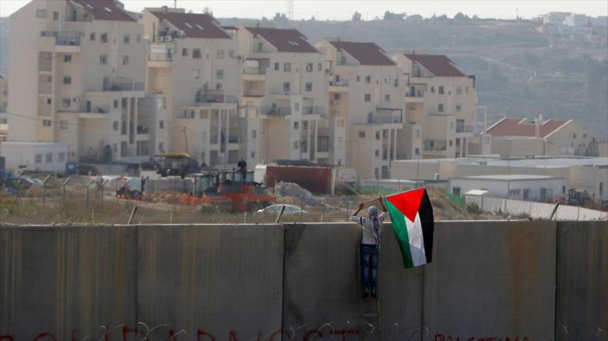 Un hombre coloca la bandera palestina en el polémico muro de separación israelí, con asentamientos ilegales judíos al fondo. (Foto: Reuters)