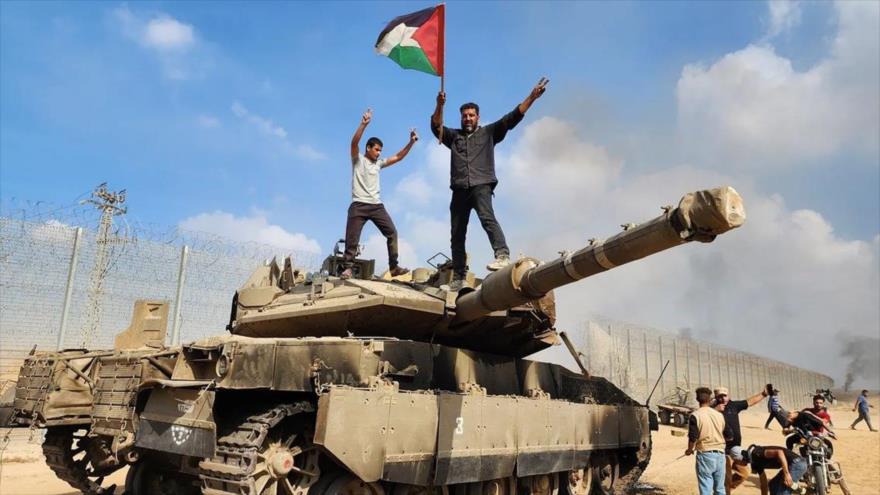 Palestinos sostienen una bandera palestina encima de un tanque israelí destruido durante la operación Tormenta de Al-Aqsa, 7 de octubre de 2023.