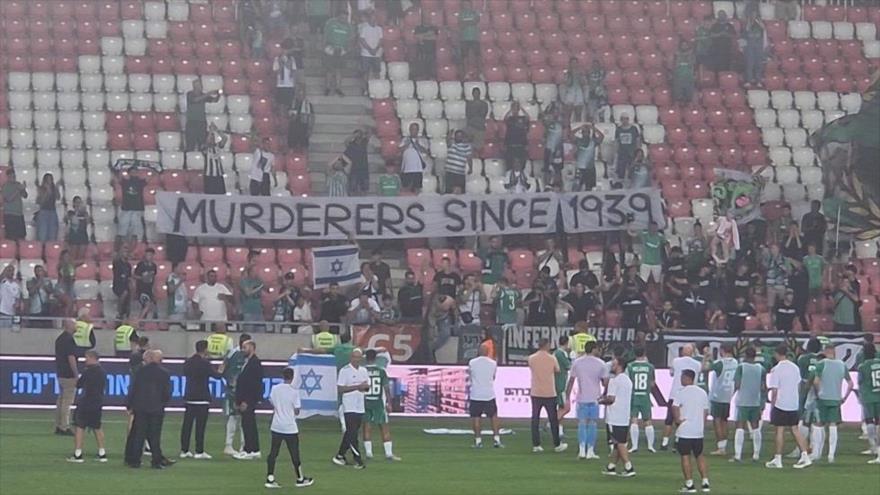 Aficionados del Maccabi Haifa muestran una pancarta con que dice “Asesinos desde 1939” durante un partido contra el club polaco Raków Częstochowa, 15 de agosto de 2025. (Foto: redes sociales).