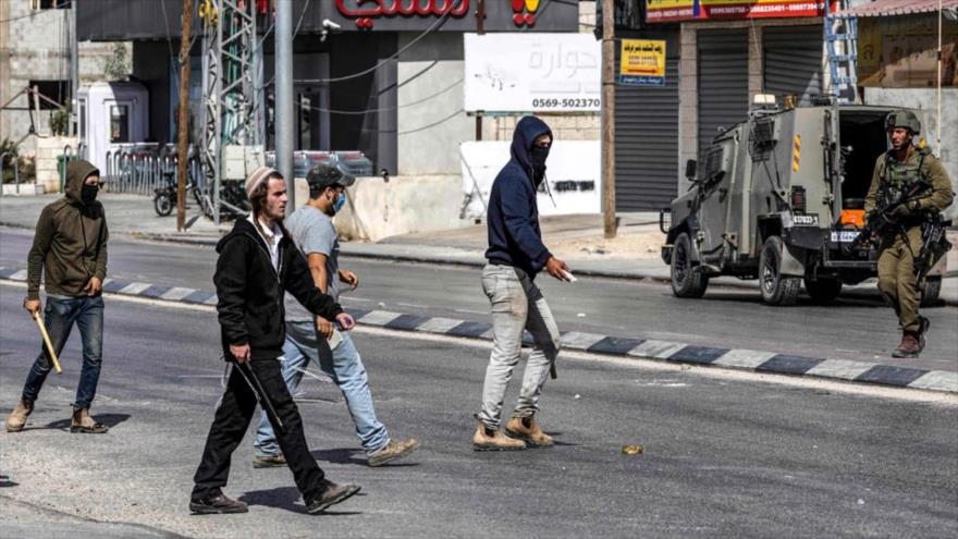Colonos israelíes caminan con palos junto a soldados sionistas durante enfrentamientos en los que atacaron a residentes y comercios palestinos en la localidad de Huwara, en Cisjordania ocupada. (Foto: AFP)